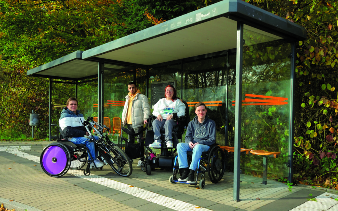4 Auszubildende des Berufsbildungswerks Volmarstein, mit Rollstuhl und Gehhilfe, an einer Bushaltestelle.
